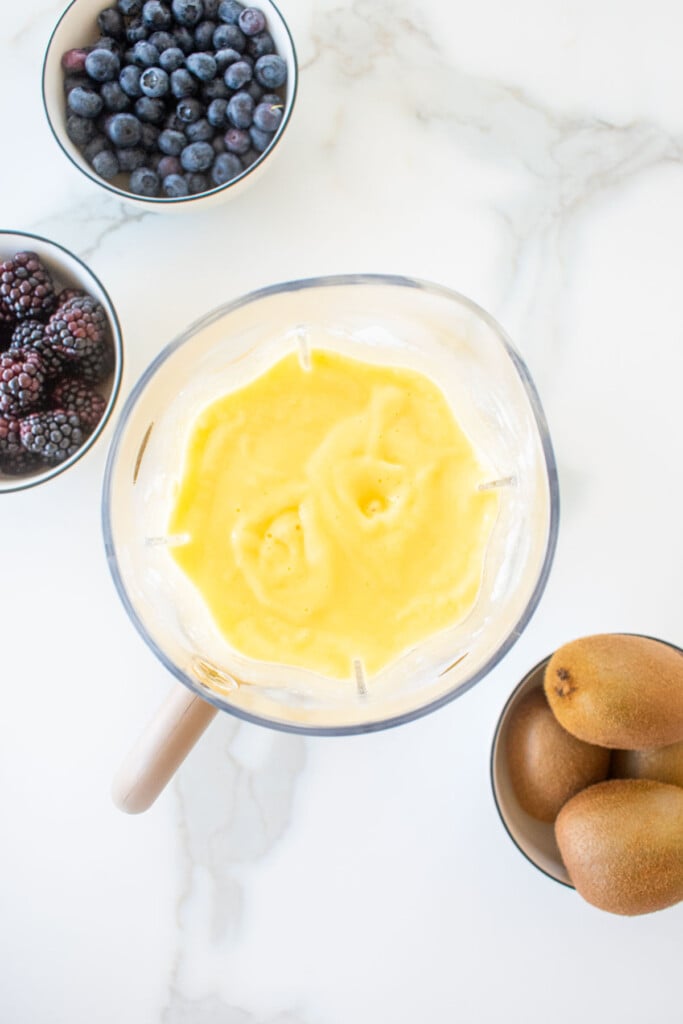 mango pineapple smoothie blended until smooth in a blender on a marble counter with fresh fruit in bowls beside it.