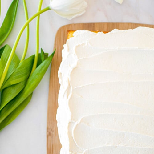 whipped cream frosting on a vanilla sheet cake on a bamboo board on a white marble counter with white tulips.