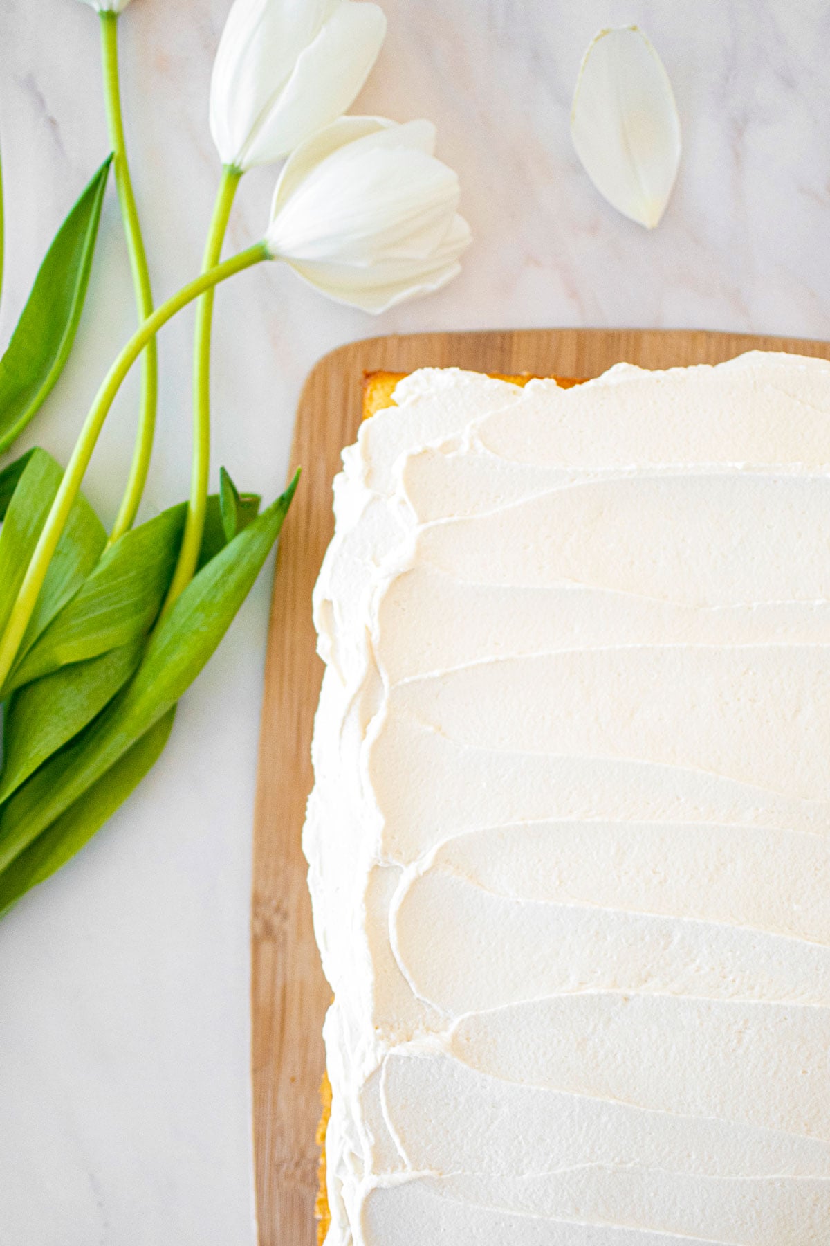 whipped cream frosting on a vanilla sheet cake on a bamboo board on a white marble counter with white tulips.
