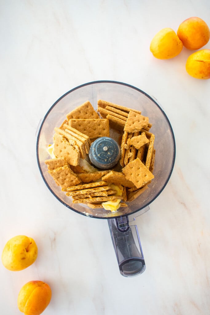 food processor with graham crackers, cane sugar and butter on a marble counter with apricots.