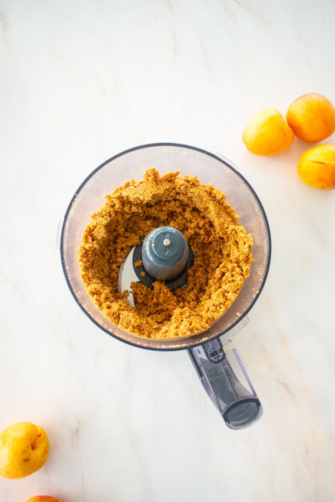 food processor with graham cracker crust blended on a white marble counter with apricos.