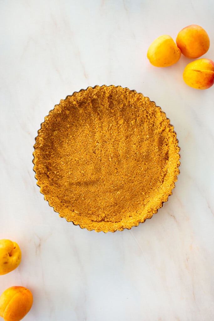 graham cracker crust in a tart pan on a marble counter with apricots.