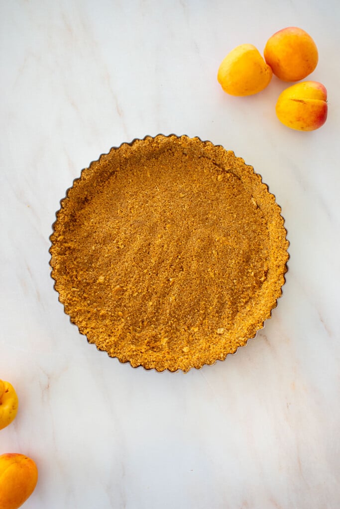 baked graham cracker crust in a tart pan on a marble counter with apricots.