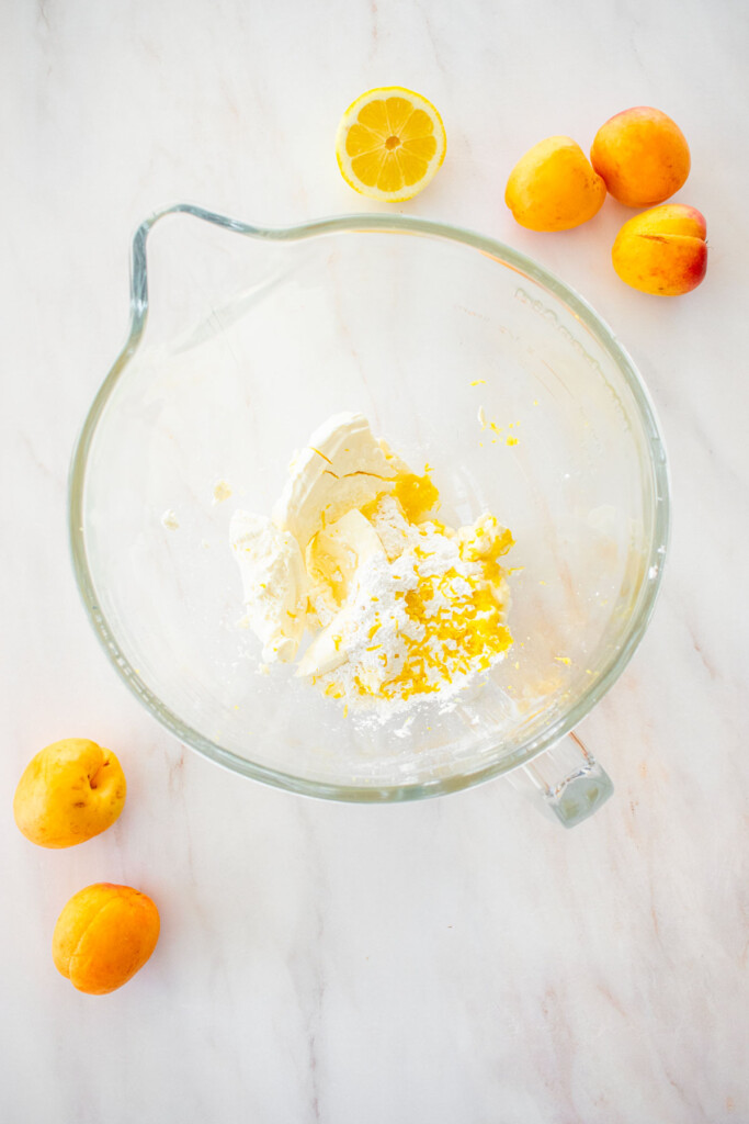 glass mixing bowl with cheesecake filling ingredients on a marble counter with fresh apricots and lemon.