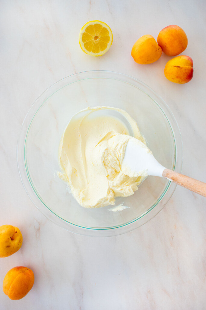 glass mixing bowl with smooth and creamy cheesecake filling and a spatula on a marble counter with fresh apricots and lemon.