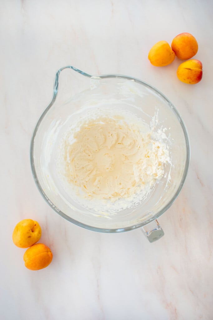 glass mixing bowl with whipped cream on a marble counter with fresh apricots.