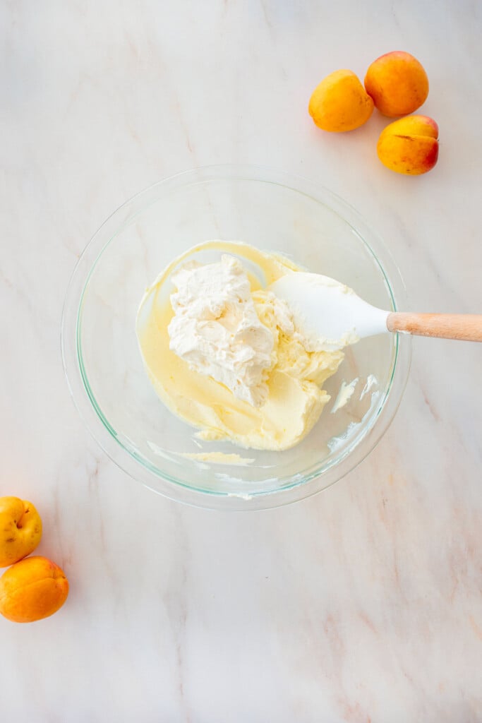 glass bowl with creamy cheesecake filling and whipped cream on top on a marble counter with fresh apricots.