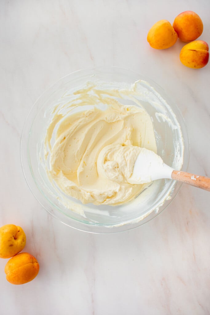 glass bowl with creamy cheesecake filling with a spatula on a marble counter with apricots.