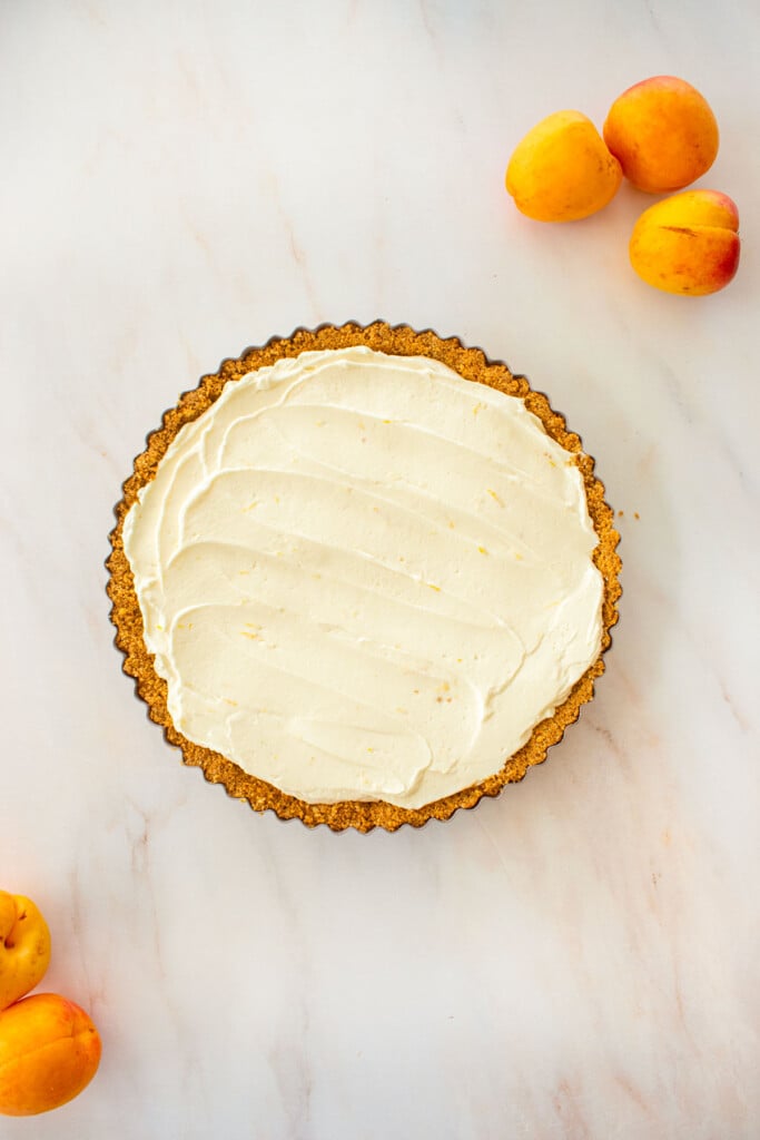 graham cracker crust in a tart pan with cheesecake filling spread evenly on a marble counter with apricots.