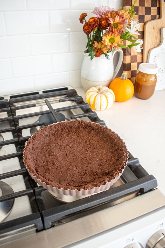 chocolate graham cracker crust in a tart pan cooling on top of an oven.