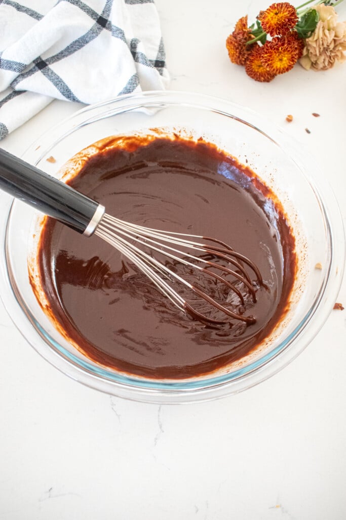 glass mixing bowl with chocolate ganache being stirred with a whisk on a marble counter with fresh flowers.
