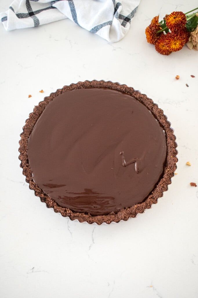 chocolate graham cracker crust with chocolate ganache spread evenly inside sitting on top of a marble counter with fresh flowers.