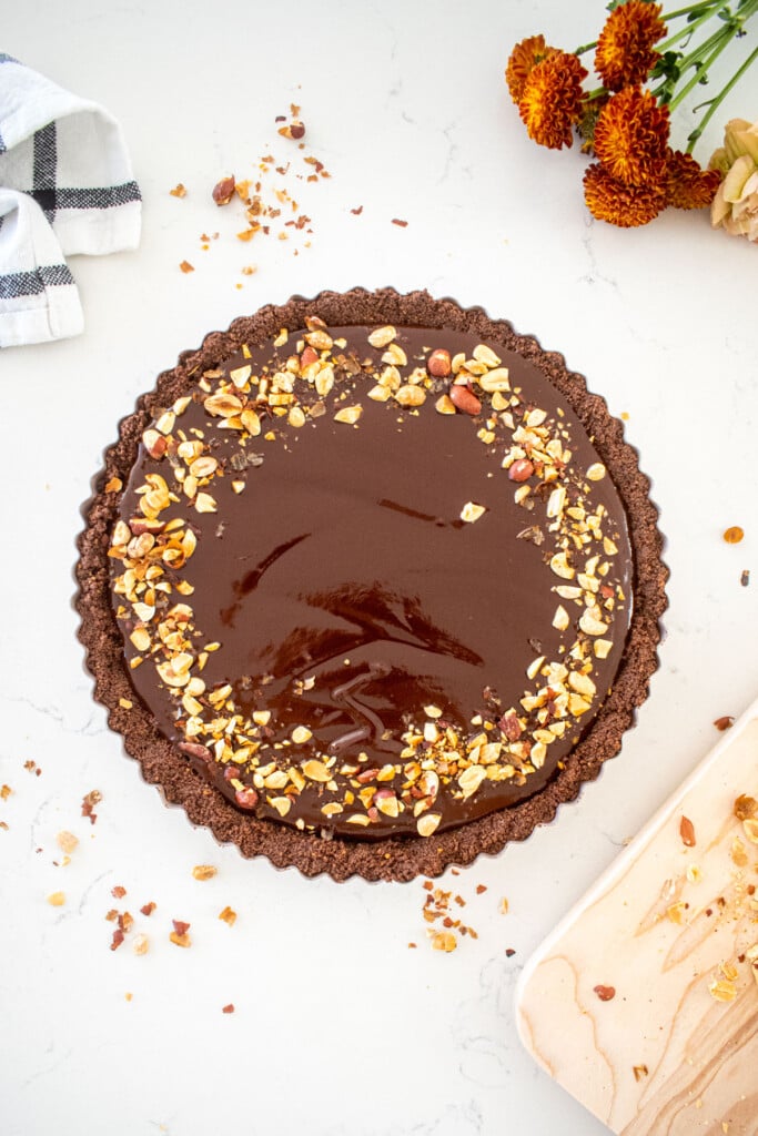 chocolate peanut butter tart with chopped peanuts on top sitting on a marble countertop with fresh flowers.