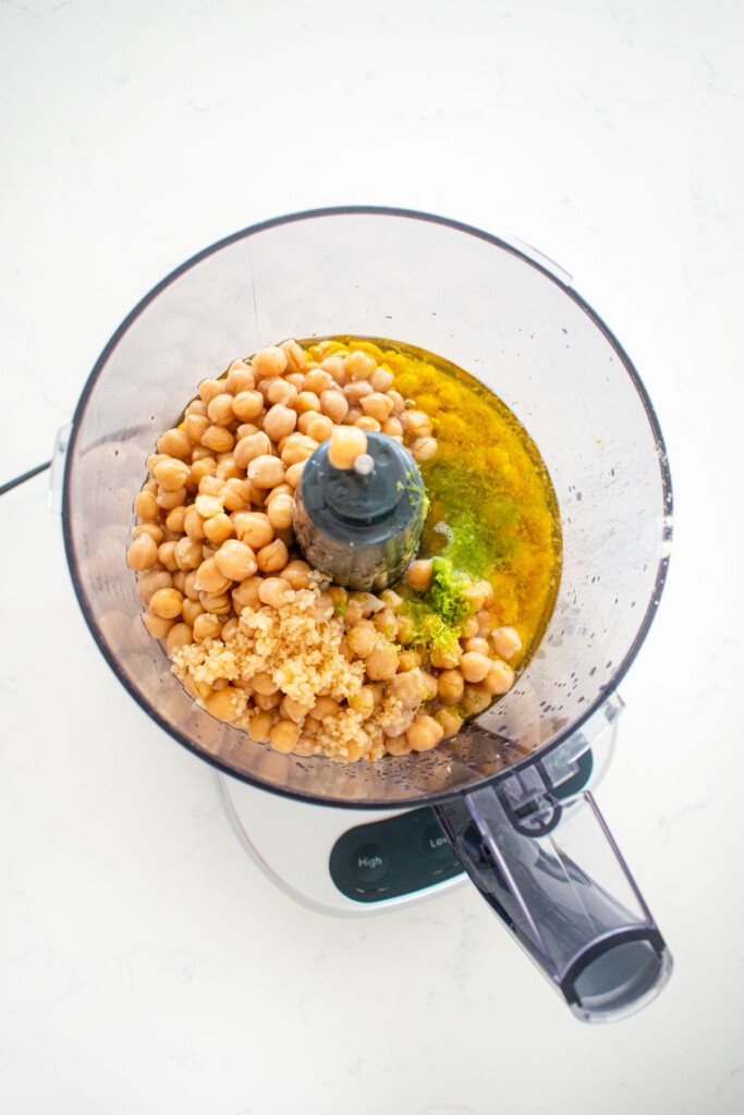 ingredients for garlic lime hummus in a food processor on a white marble countertop.