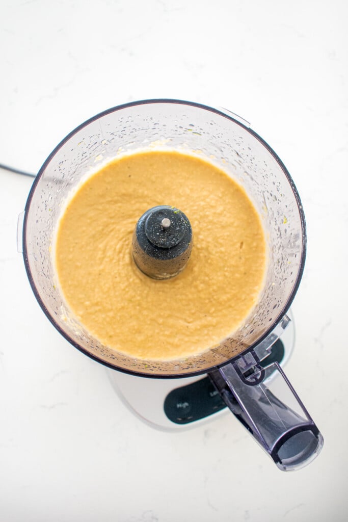 blended garlic lime hummus in a food processor on a white marble countertop.