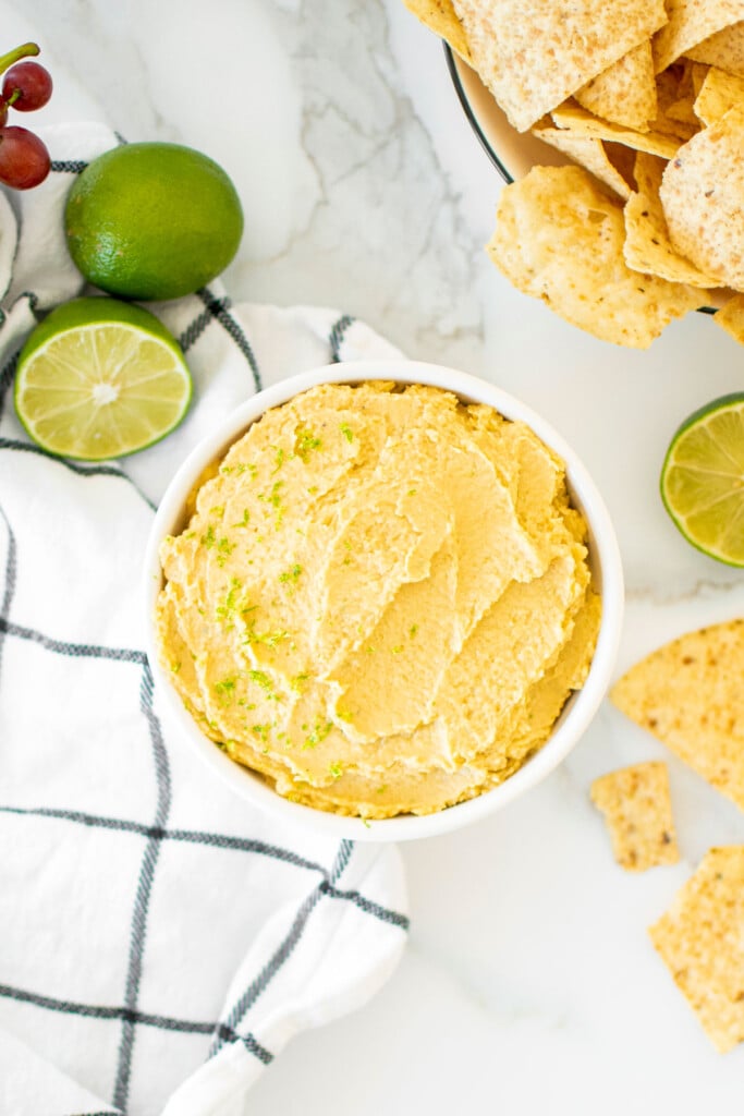 garlic lime hummus in a bowl on a white marble countertop with tortilla chips and fresh limes.