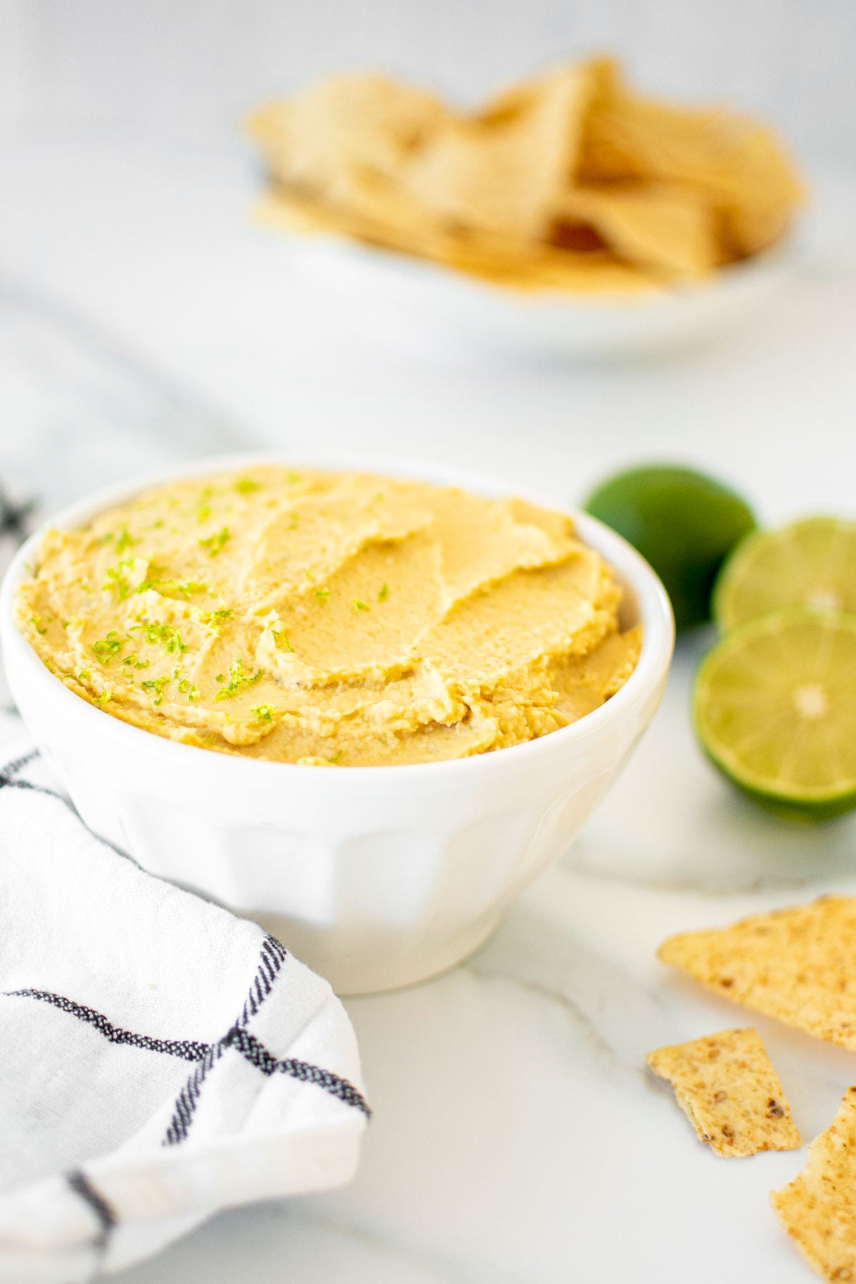 garlic lime hummus in a bowl on a white marble countertop with tortilla chips and fresh limes.