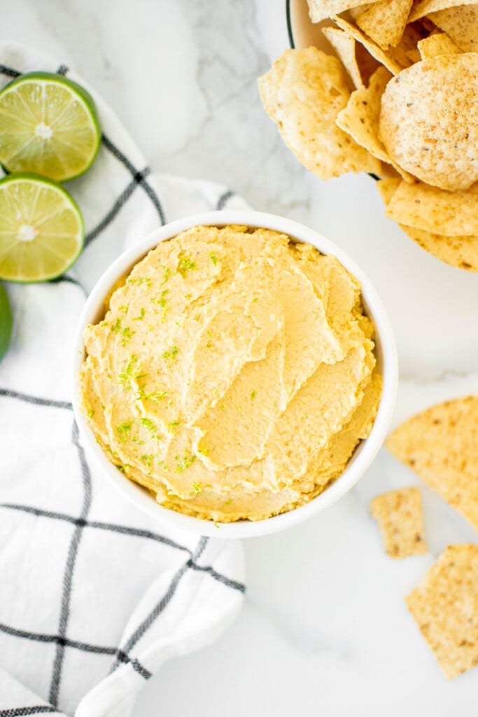garlic lime hummus in a bowl on a white marble countertop with tortilla chips and fresh limes.