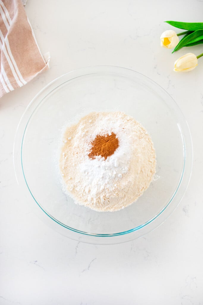 large glass mixing bowl with flour, baking powder, salt and cinnamon on a white marble counter.