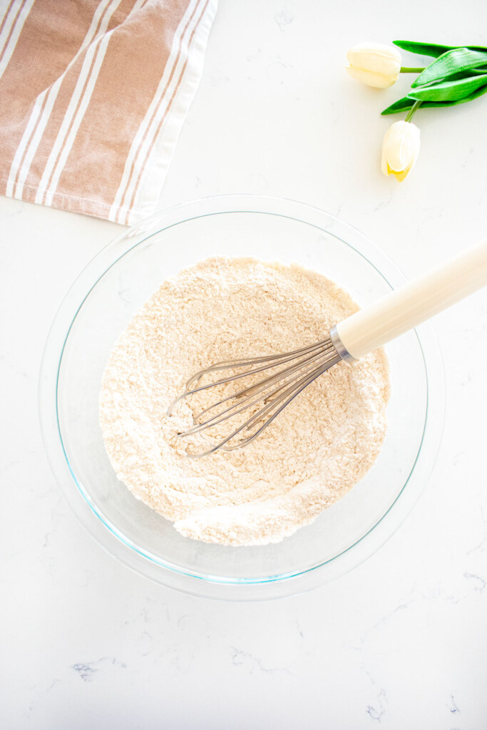 large glass mixing bowl with flour, baking powder, salt and cinnamon being whisked together on a white marble counter.
