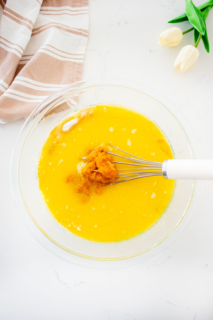 eggless waffle batter being mixed with pumpkin puree in a glass mixing bowl with a whisk on a white marble counter.