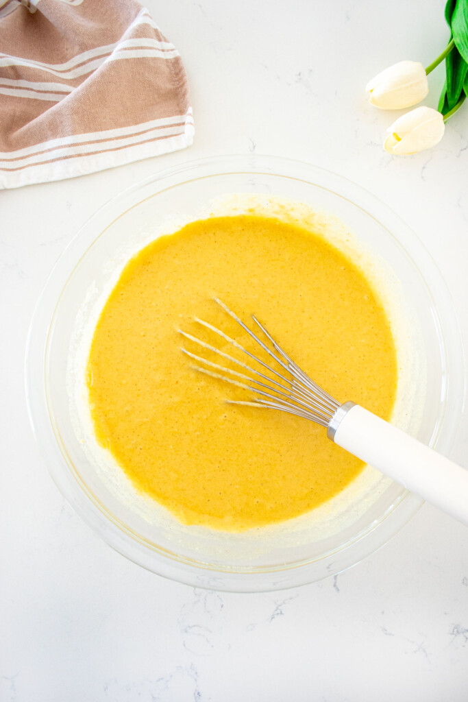eggless waffle batter in a glass mixing bowl with a whisk on a white marble counter.