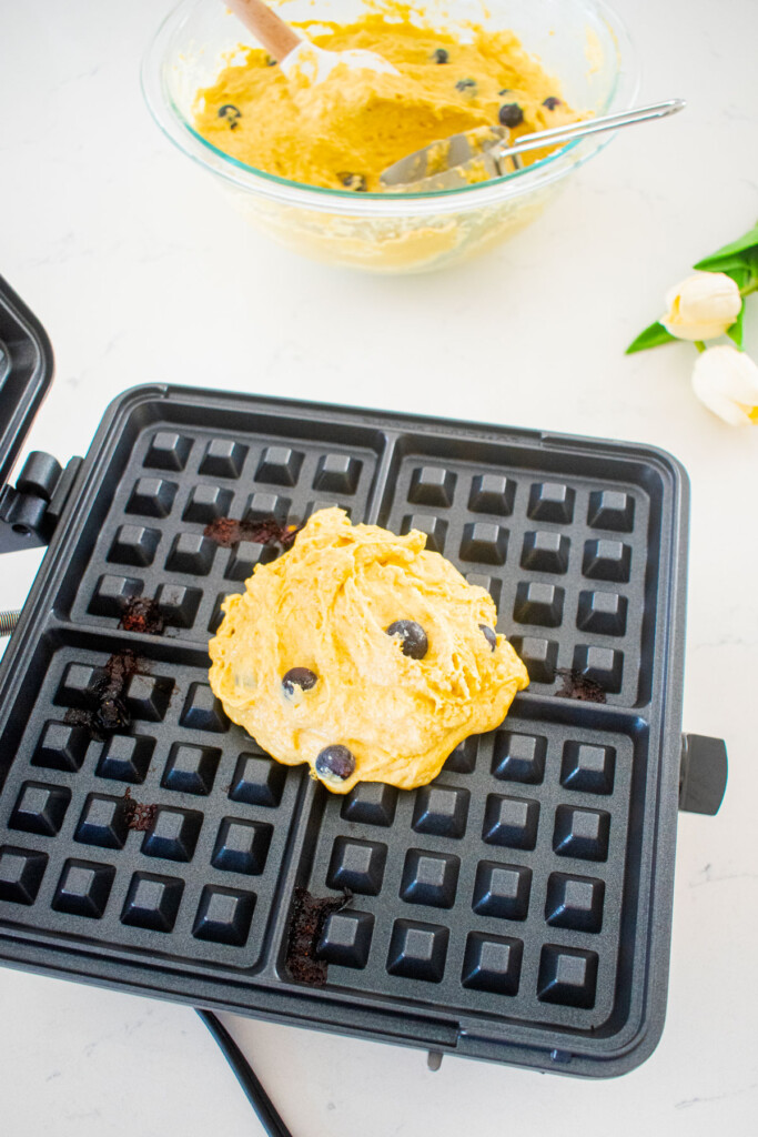 eggless blueberry waffle batter in a waffle iron on a white marble counter.