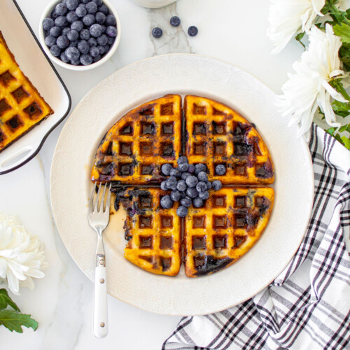 homemade blueberry waffles without eggs on a plate on a white marble counter with fresh blueberries and maple syrup.