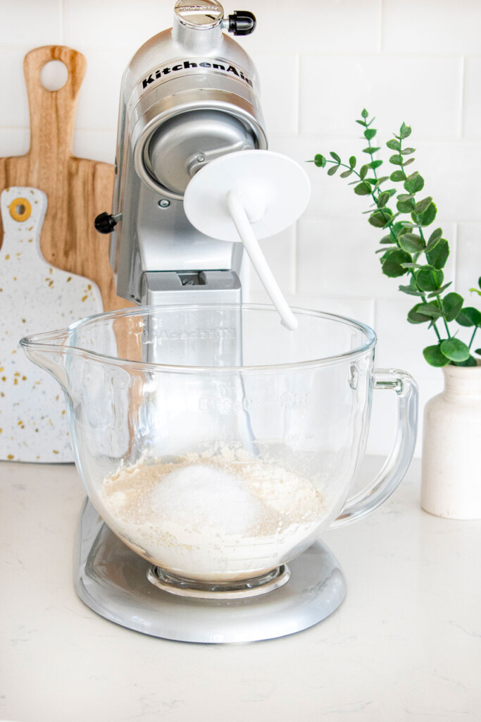 ingredients for brioche dough in a glass bowl of a stand mixer on a white kitchen countertop.