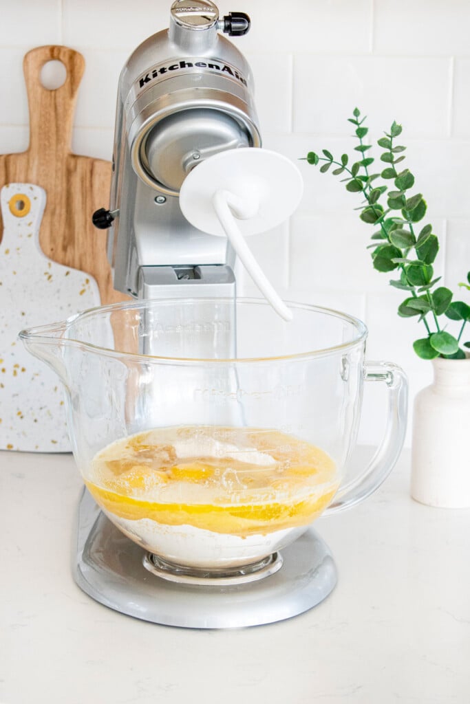 ingredients for brioche dough in a glass bowl of a stand mixer on a white kitchen countertop.