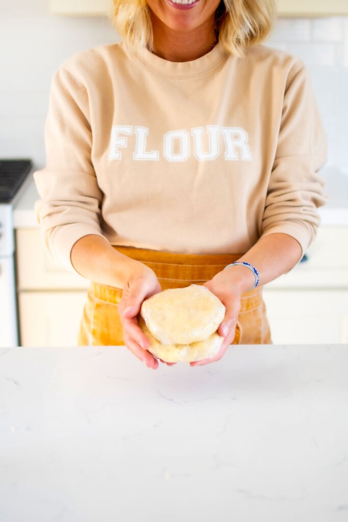 woman holding pie crust wrapped in plastic wrap at kitchen counter wearing a flour sweatshirt and apron.