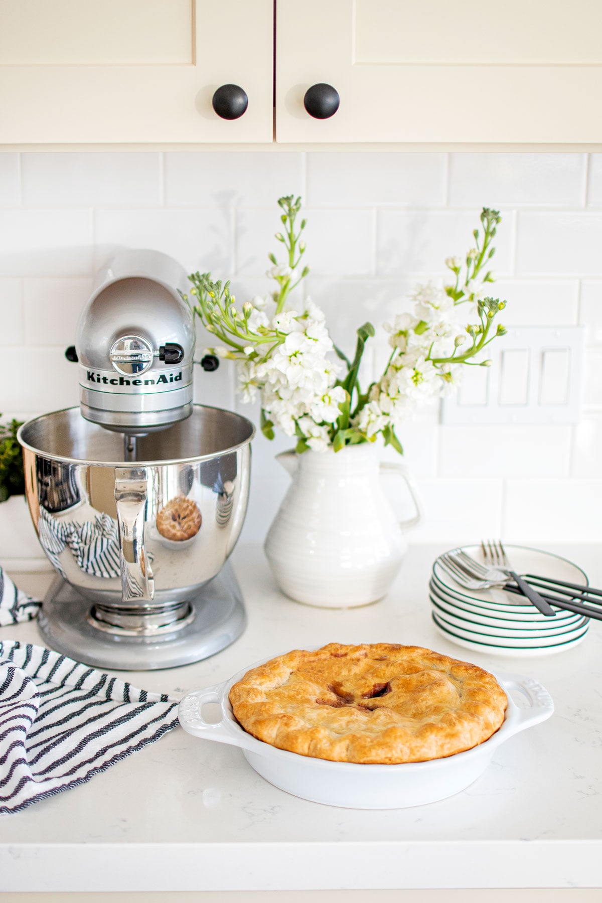 berry pie in a white pie pan on a marble counter with a stand mixer and vase of flowers and dessert plates.