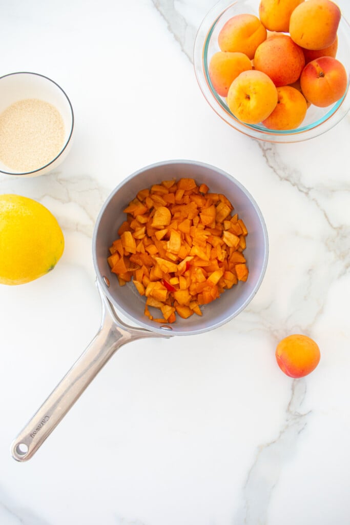 chopped apricots in a small pot on a marble counter with a bowl of apricots, cane sugar and lemon on the side.