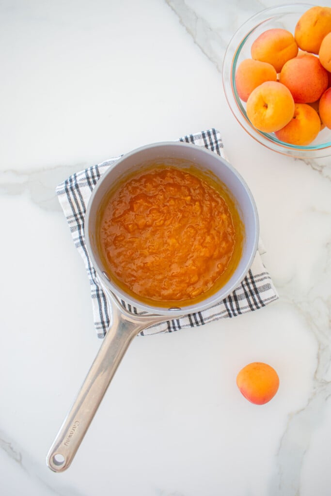 apricot jam in a small pot on a kitchen towel on a marble counter.