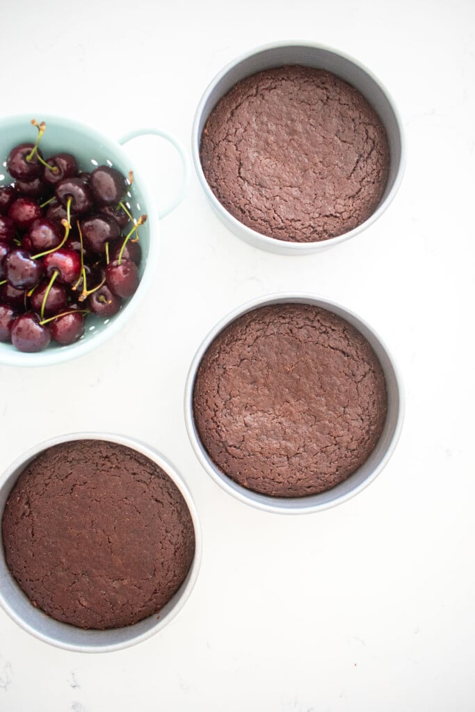baked chocolate cake in three 6-inch cake pans on a white marble counter with a bowl of cherries.