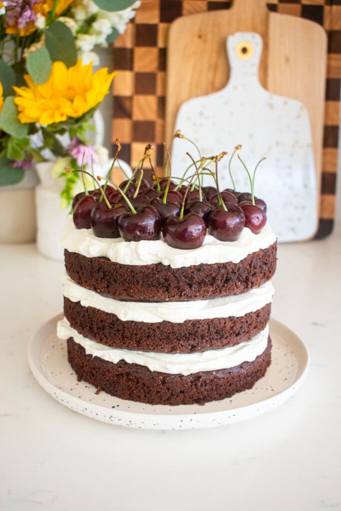 chocolate cherry whipped cream cake with fresh cherries on top on a white marble counter with fresh flowers.