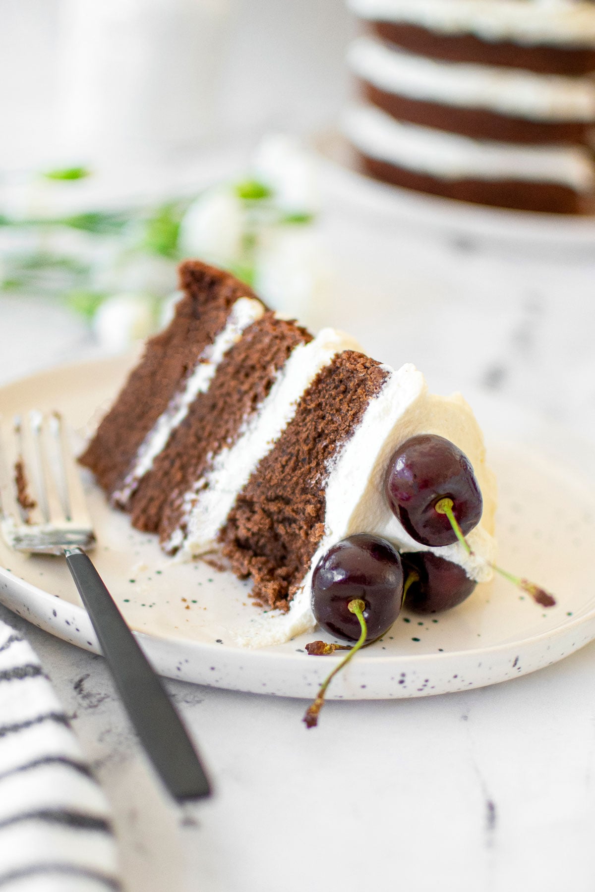 chocolate cherry whipped cream cake slice on a plate with a fork on a marble table.