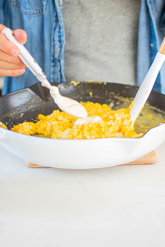 woman adding sour cream to scrambled eggs in a white cast iron skillet on a white marble counter.