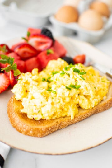 fluffy scrambled eggs with sour cream on top of sourdough toast with fresh chives and fresh fruit on a plate.