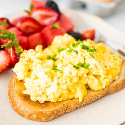 fluffy scrambled eggs with sour cream on top of sourdough toast with fresh chives and fresh fruit on a plate.