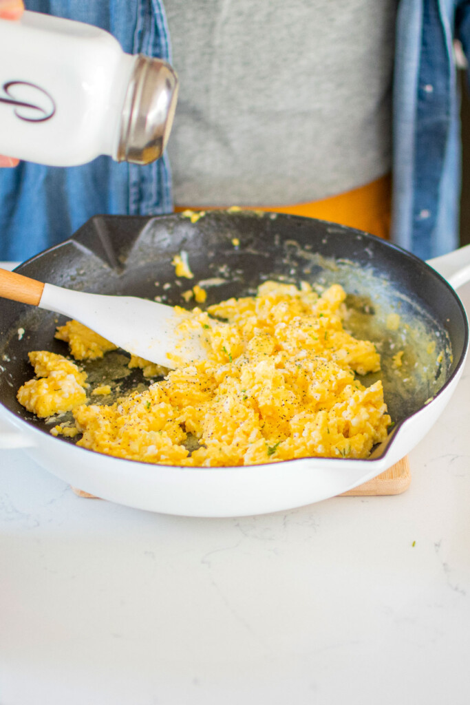 woman salting fluffy scrambled eggs with sour cream in a white cast iron skillet on a marble counter.