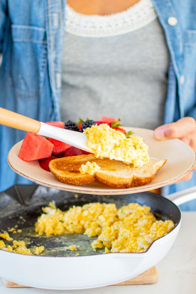 woman serving fluffy scrambled eggs on top of toast on a plate with fresh fruit.