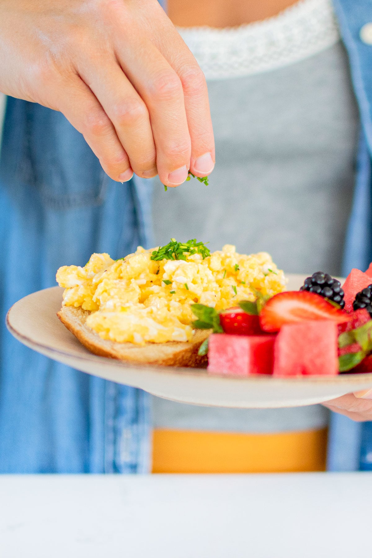 woman adding fresh chives on top of fluffy scrambled eggs with sour cream on a plate with fresh fruit.