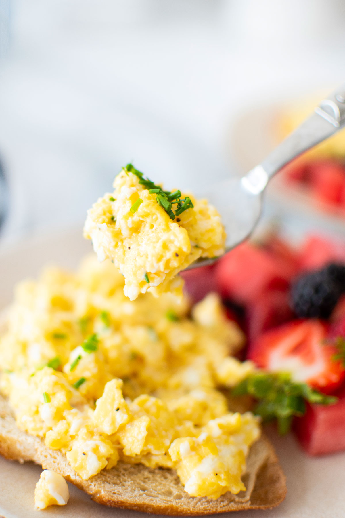 fluffy scrambled eggs on a fork with fresh chives being scooped off a plate with eggs and fresh fruit.