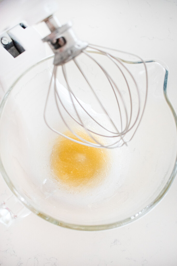 gelatin and water blooming in a glass mixing bowl with a whisk on a white marble counter.