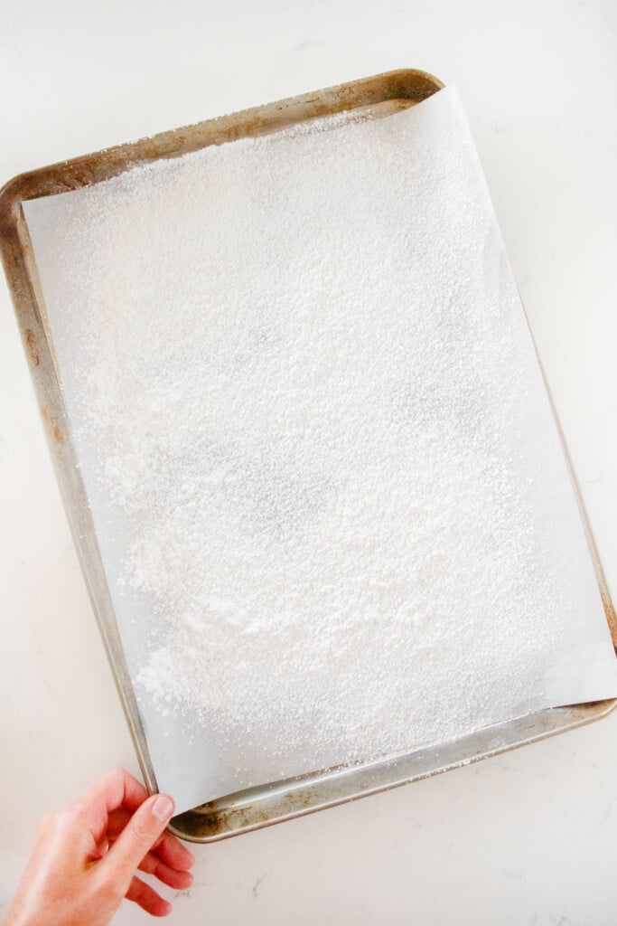 a half sheet pan with parchment paper and powdered sugar sifted on top of it on a white marble counter.