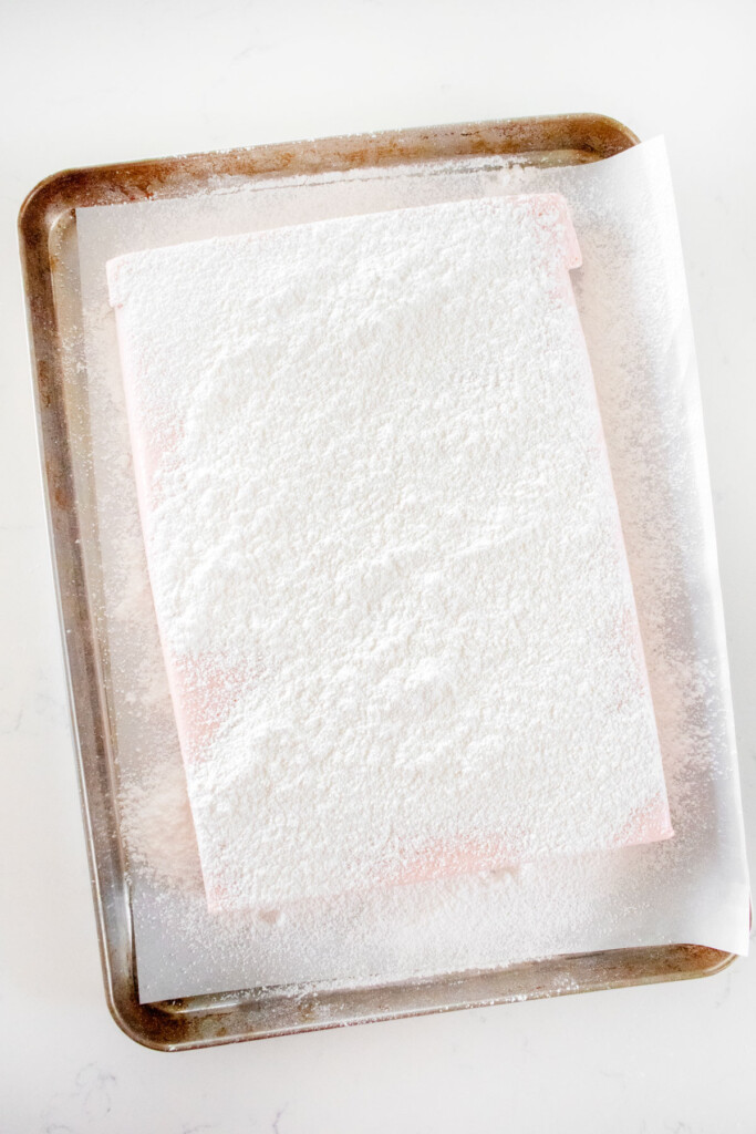 homemade cherry marshmallows on a baking sheet with sifted powdered sugar on top on a white marble counter.