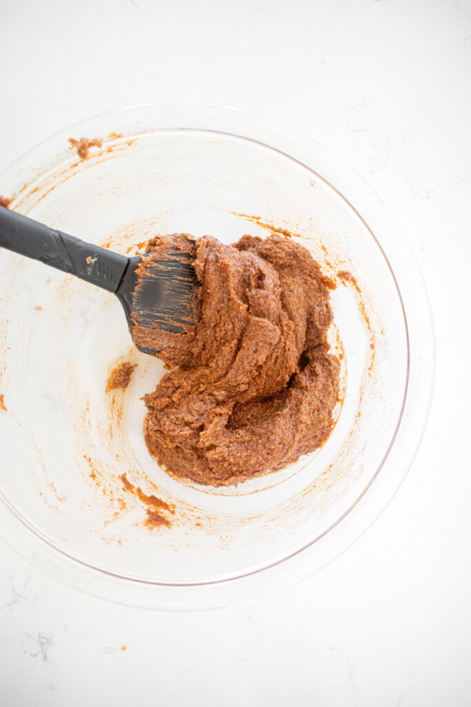cinnamon roll filling mixed together with a spatula in a glass mixing bowl on a white marble counter.