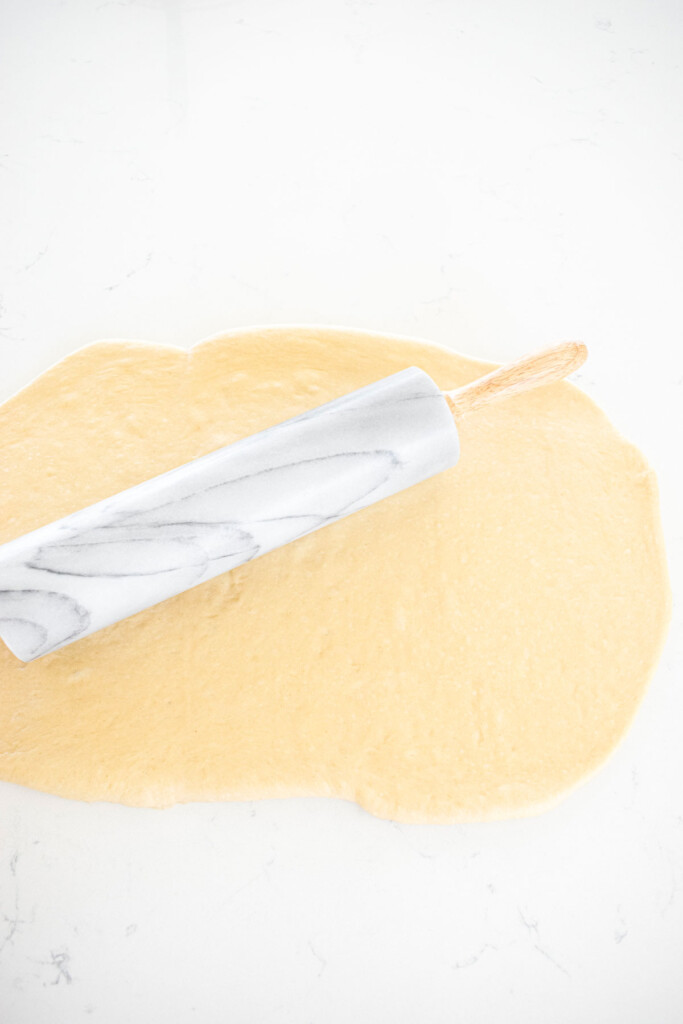cinnamon roll dough being rolled out with a rolling pin on a white marble counter.