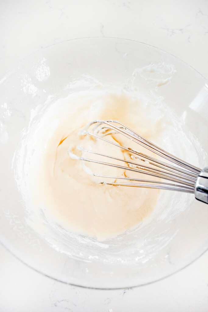 glaze with a whisk in a glass mixing bowl on a white marble counter.